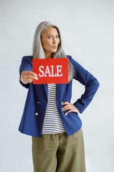 Cute mature Asian lady with loose grey hair wearing elegant jacket holds red card with word Sale posing on light background in studio