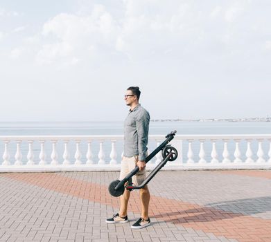 Young man holding folding electric scooter on city embankment in front of sea.