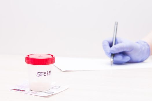 a test jar with a red cap and the inscription sperm costs a banknote of 500 euros, against the background a hand in a blue disposable medical glove writes with a pen on white paper, copy space, sperm in a jar, sperm donor concept