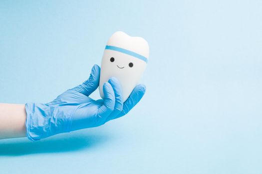 hand in a blue disposable rubber medical glove holds a toy smiling tooth on a blue background copy space, pediatric dentistry concept, treatment of a toothache