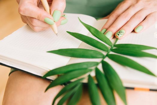 Unrecognizable young woman with notebook, a pen and green palm leaf. Girl writing romantic notes.