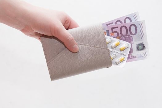 female hand holds a wallet with yellow capsule pills and euro notes, white background