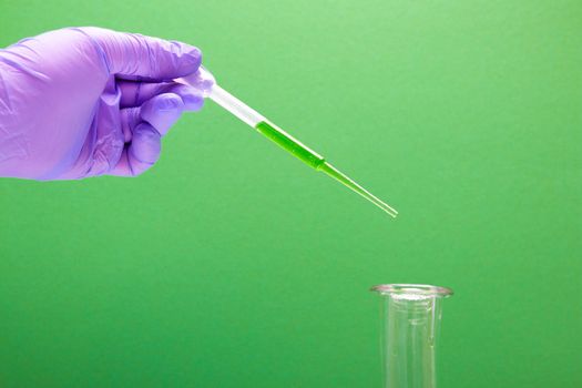 a female hand in a blue disposable glove holds a large plastic dropper, drips green liquid into a flask, green background, copy space
