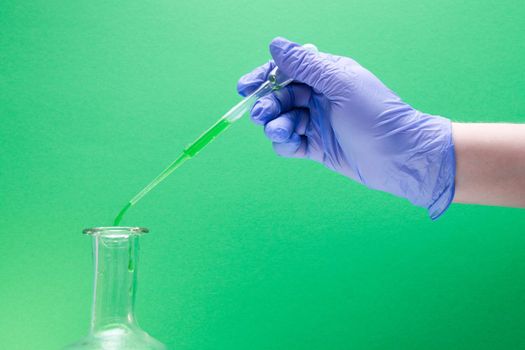 a female hand in a blue disposable glove holds a large plastic dropper, drips green liquid into a flask, green background, copy space