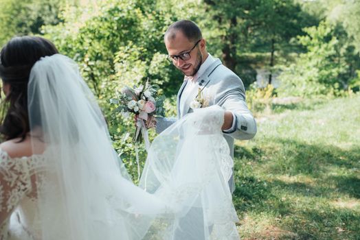 Bearded groom in a gray jacket and glasses holding a veil or wedding dress of the bride.
