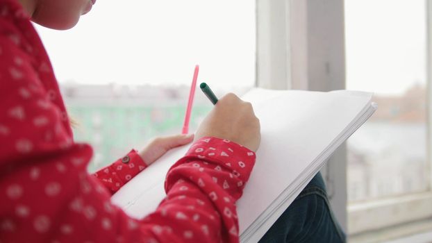 A little girl sitting on the window sill and drawing, smiling on the backround of the city. Close up photo. Portrait