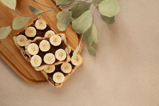 Three banana white bread toasts smeared with chocolate butter that lie on a cutting board with a sprig of leaves on craft paper background. top view with area for text