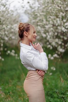 Beautiful young blonde woman in white shirt posing under apple tree in blossom and green grass in Spring garden