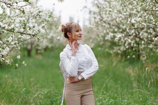 Beautiful young blonde woman in white shirt with backpack posing under apple tree in blossom and green grass in Spring garden