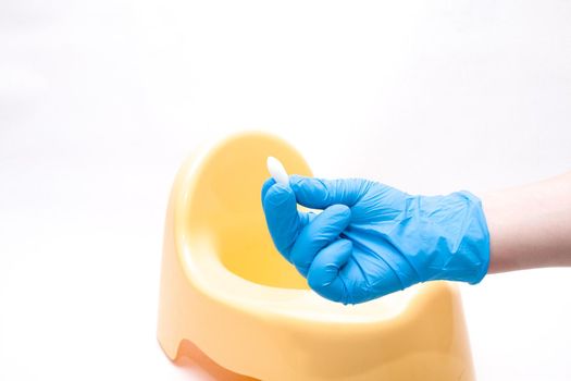 a hand in a disposable blue medical glove holds a test jar with a white spoon for collecting feces, in the background is a yellow children's pot, white background, copy space