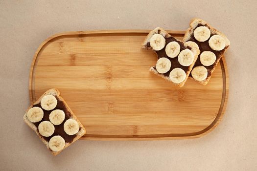 Three banana white bread toasts smeared with chocolate butter that lie on a cutting board on craft paper background. top view with area for text