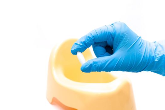 a hand in a disposable blue medical glove holds a test jar with a white spoon for collecting feces, in the background is a yellow children's pot, white background, copy space