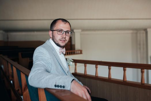 Wedding photo of emotions of a bearded groom with glasses in a gray jacket in the church building.