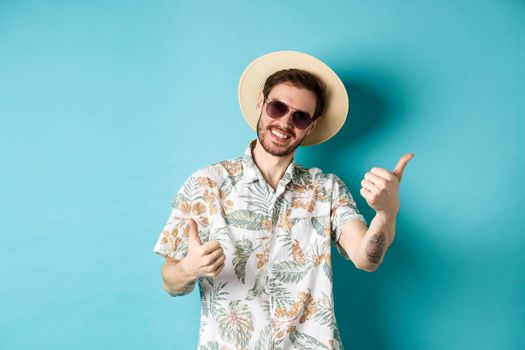 Cheerful tourist having fun summer holidays, showing thumbs up and smiling, standing in hawaiian shirt and sunglasses on blue background.