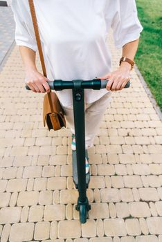 Unrecognizable woman in white clothing riding an electric scooter in summer park, front view.