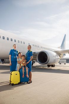 Full length shot of beautiful stewardesses standing near two kids sitting on their luggage in front of big airplane and smiling at camera. Family, traveling, vacation concept