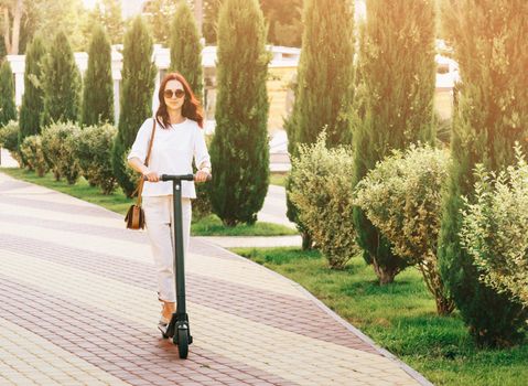 Stylish beautiful young woman in white clothing riding an electric scooter in city park on sunny summer day.