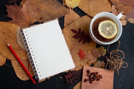 red pencil, notebook, raisins, a cup with lemon tea lie on autumn dry leaves on a black table background top view flat lay