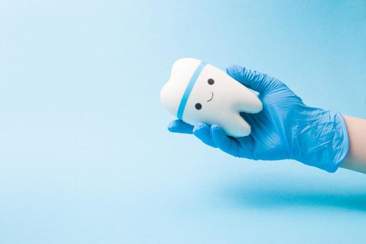 hand in a blue disposable rubber medical glove holds a toy smiling tooth on a blue background copy space, pediatric dentistry concept, treatment of a toothache