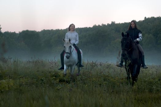Two women on a foggy field riding horses towards the camera. Wide shot
