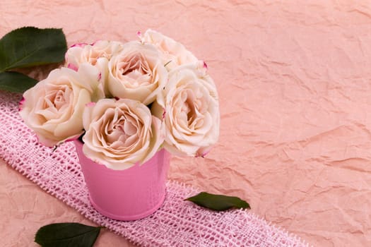 A bouquet of beautiful roses stands in a small bucket on a lace ribbon on a pink craft background with space for text.