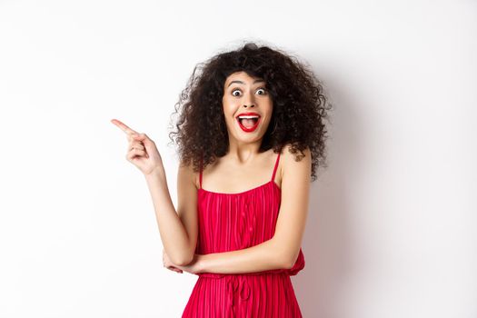 Excited smiling woman with curly hair and makeup, wearing elegant red dress, scream surprised and pointing finger left at logo, standing on white background.