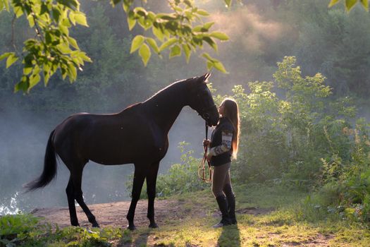 Young woman with her brown horse on a beautiful landscape. Clear lake at morning fog. Sunrise. Wide shot