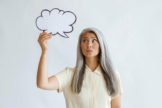 Doubting mature woman with long grey hair holds blank text bubble standing in light background in sudio. Mockup for design