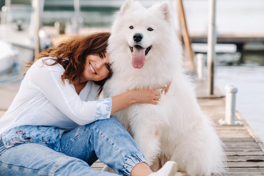 a happy woman with a big white dog sits on a pier by the sea at sunset.