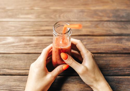 Woman’s hands with bright orange manicure holding bottle of fresh berry smoothie at wooden table. Point of view in first person.