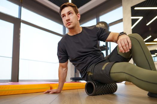 The man is massaging his leg muscles with the foam roller in gym