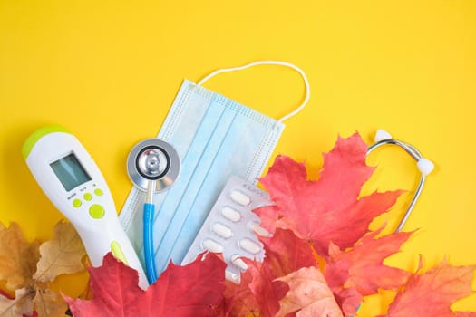 non-contact thermometer, drugs, disposable protective medical mask and stethoscope and autumn maple leaves on a yellow background copy space covid 19