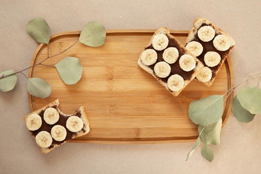 Three banana white bread toasts smeared with chocolate butter that lie on a cutting board with a sprig of leaves on craft paper background. top view with area for text