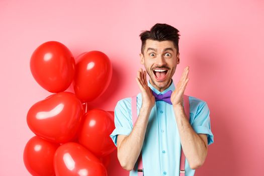 Love and romance concept. Happy man screaming from fantastic news, shouting wow and smiling amused, checking out special offer on Valentines, standing near red hearts balloons.