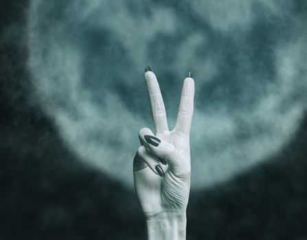 Hand of a dead zombie man with peace sign gesture in front of moon. Halloween and horror concept.