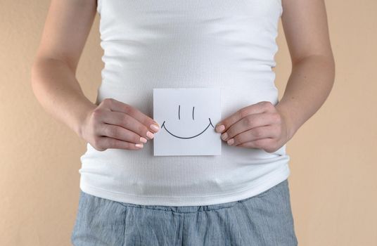 A close-up view of the belly of a pregnant woman in a white T-shirt that is holding a piece of paper with a smiley face
