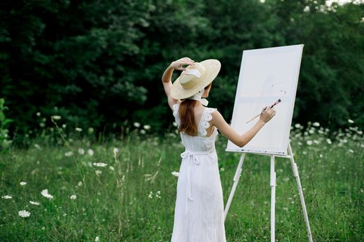 A woman in a white dress in a field with flowers paints a picture. High quality photo