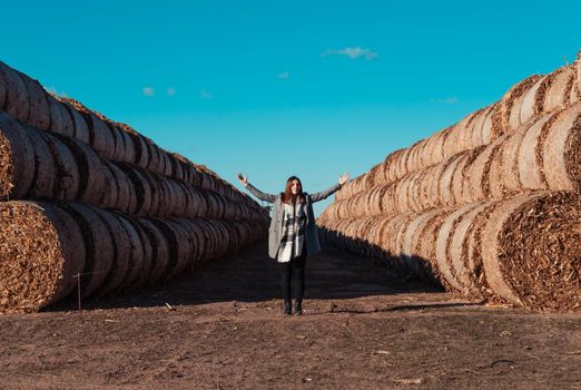 A girl of European appearance in a gray coat stands in a field near a larger bale with hay against a blue sky