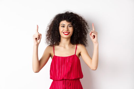 Smiling woman in elegant red dress and makeup, pointing fingers up and showing promo offer on valentines day, standing over white background.