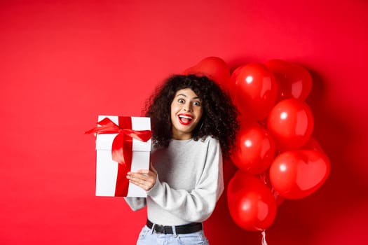 Surprised and happy woman holding valentines day gift from lover, standing near romantic hearts balloons and looking at camera amazed, red background.