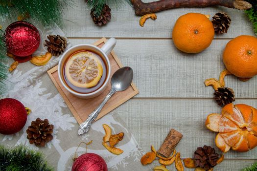 cup of tea with lemon new year's decor on a wooden background gray table, green pine branch, tangerines, spoon, dried fruits, christmas balls, top view, copy space