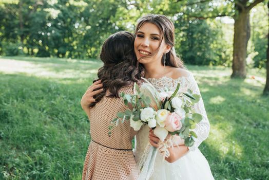 Beautiful bride in a wedding dress in the forest is hugging a girl.