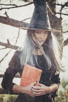 Dark young witch standing with book in the forest, looking at camera. Space for text on book. Vintage image