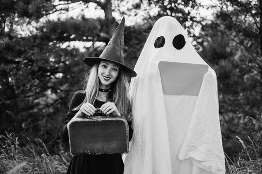 Funny Halloween couple: Cheerful witch with suitcase and white ghost with empty poster in autumn forest, space for text. Monochrome image