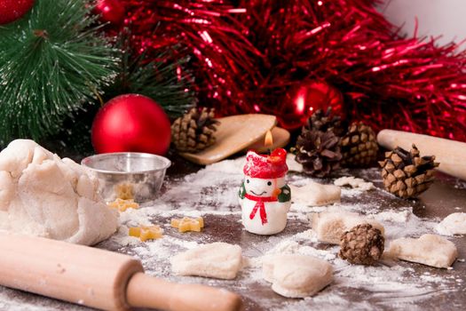 cooking christmas baking, christmas background, dough, kitchen creature, wooden table, a burning snowman candle, a branch of sona, various cones and decorations in the background