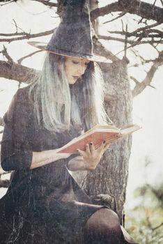 Beautiful young witch sitting on old dry tree and reading a book of magic outdoor. Vintage image