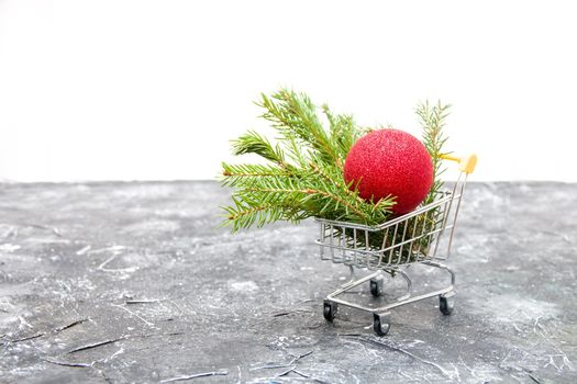 spruce green branch and red shiny Christmas-tree toy ball in a miniature shopping trolley on a black background, copy space