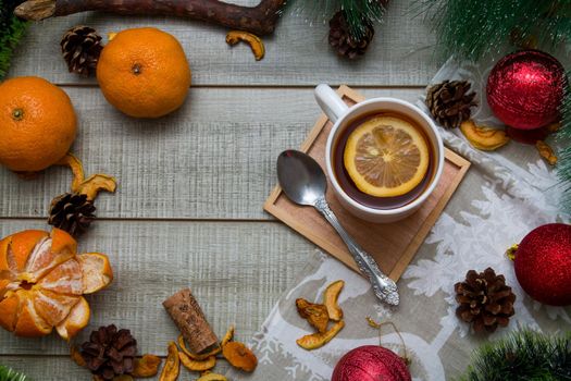 cup of tea with lemon new year's decor on a wooden background gray table, green pine branch, tangerines, spoon, dried fruits, christmas balls, top view, copy space