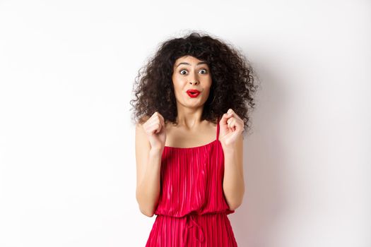 Excited smiling woman eager to try something, jumping from amusement and smiling, wearing red dress, standing on white background.