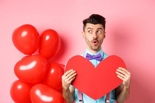 Valentines day concept. Cute young man looking left amused, showing red heart cutout and standing near balloons, pink background.